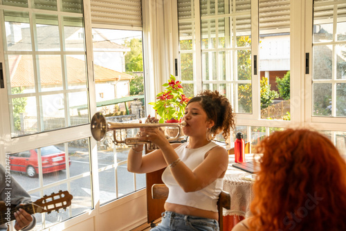Sticker Curly haired young woman plays trumpet indoors beside large windows, with sunlig