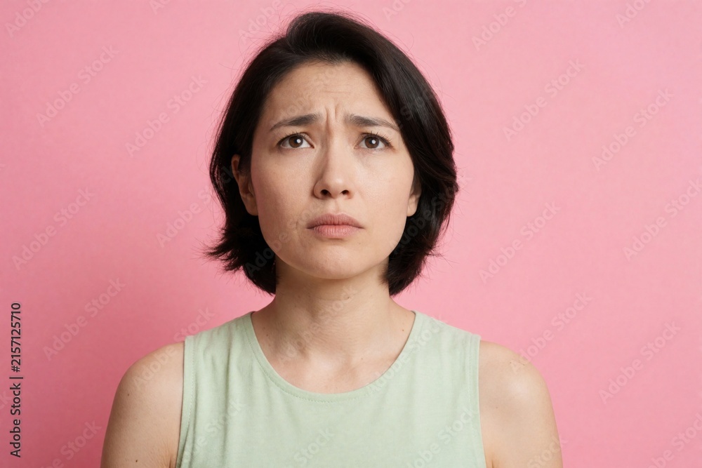 Fototapeta premium Young woman frowns looking up with worried expression, biting her lip. Female has concerned face, troubled thoughts, deep thinking, negative emotion, indoor studio shot.