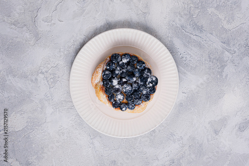 Top view of golden puff pastry Danish topped with fresh blueberries and powdered sugar on white ribbed plate, textured light gray background, copy space