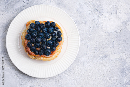 Top view of golden puff pastry Danish topped with fresh blueberries on white ribbed plate, textured light gray background, copy space