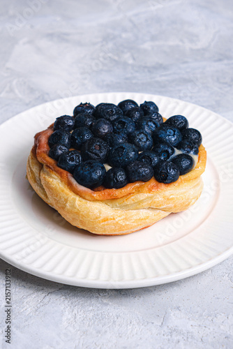 Close-up angle of golden puff pastry Danish piled with juicy fresh blueberries and cream filling on white ribbed plate, light textured background, vertical shot