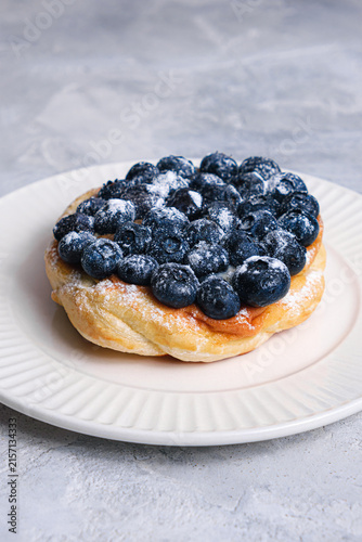 Close-up angle of golden puff pastry Danish piled with juicy fresh blueberries and cream filling on white ribbed plate, light textured background, vertical shot