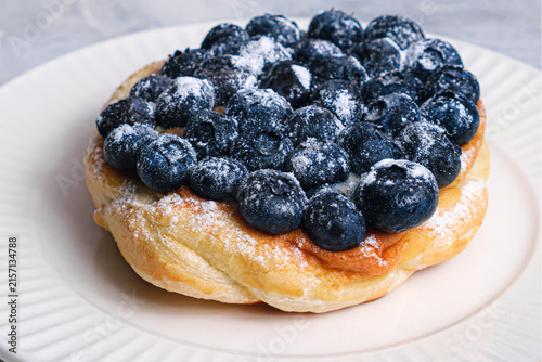 Close-up angle of golden puff pastry Danish piled with juicy fresh blueberries and cream filling on white ribbed plate, light textured background