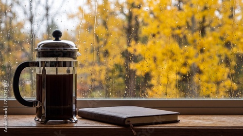 Cozy autumn morning scene with a French press coffee maker and a leather journal on a wooden windowsill against a rainy window with yellow fall foliage.