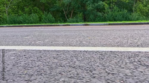 An empty asphalt road with a painted white marking line passes next to a row of green trees, the camera confidently moves forward along the asphalt surface in bright daylight. Side view