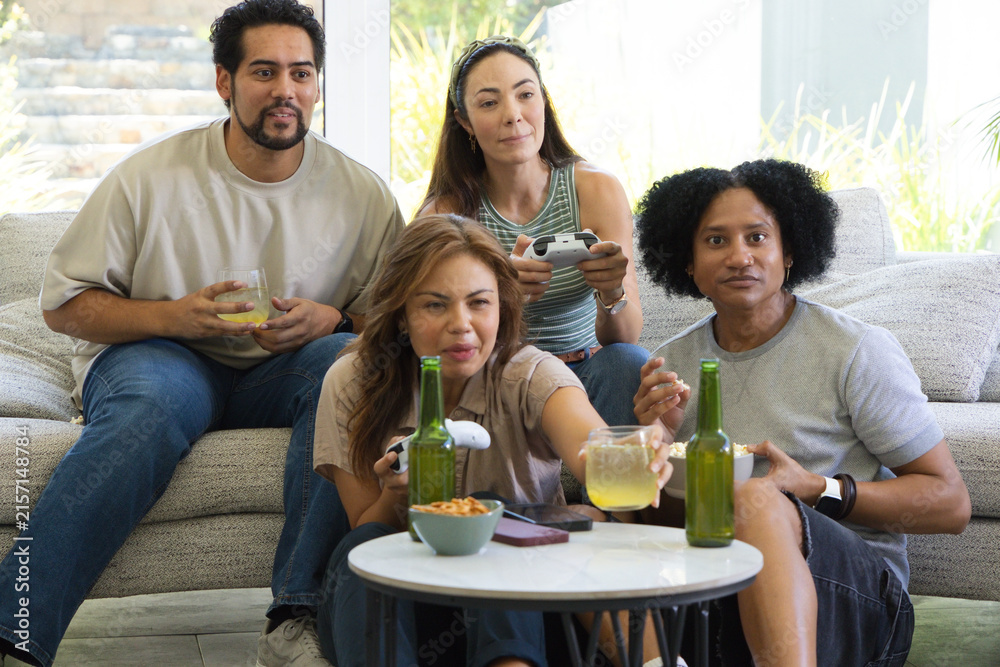 Obraz premium Diverse friends sitting on sofa and floor using game controllers at home with drinks and snacks