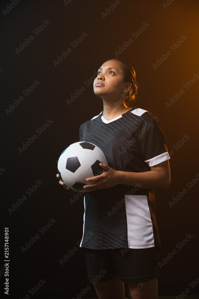 Fototapeta premium African American female soccer player holding black-white ball in black kit, studio-lit, gazing up