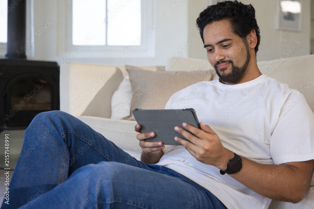 Fototapeta premium African man 30s reclining on light sofa in living room holding gray tablet wearing white tee