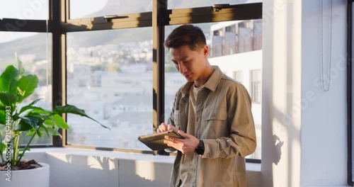 Mid Adult Asian man standing by black window, looking down at tablet, tapping, glancing up, smiling