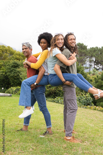 Diverse couples posing on grassy lawn in park, carrying partners while showing sunglasses, jeans