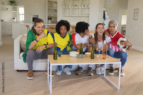Friends wearing colorful jerseys watching match on sofa at home with soccer ball beer snacks remote