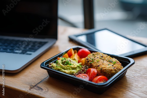 Beautifully arranged vegetarian lunch box featuring rainbow vegetables on a wooden table alongside a laptop, showcasing a fresh and balanced meal for productive days.
