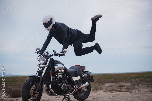 Athletic motorcyclist in white helmet performing extreme horizontal acrobatic stunt on motorcycle handlebars outdoors. Strength, freedom and extreme sports concept