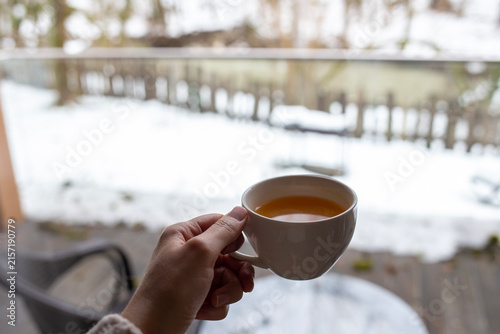 A woman's hand holding a mug of hot sea buckthorn tea while standing on a balcony of a country house, with a snowy winter forest in the background, face not visible.