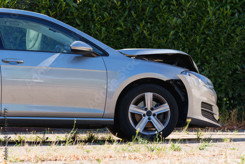 Damaged silver car with a crumpled front hood parked beside a hedge