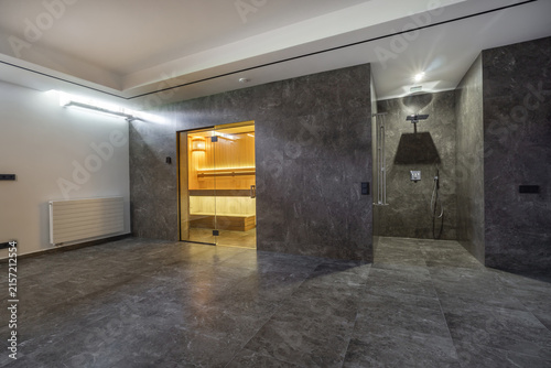 A modern spa area shows a changing room with wooden elements and a shower space. The floors are smooth and dark, while the lighting is soft and bright