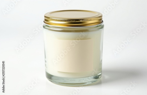 Minimalist candle in glass jar with gold lid sits on white background. Blank cream label provides branding template for small business product packaging. Transparent vessel contains white wax aroma