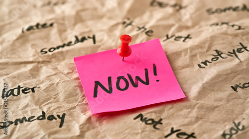 Vibrant pink sticky note with 'NOW!' message pinned to crumpled paper, surrounded by deferring words