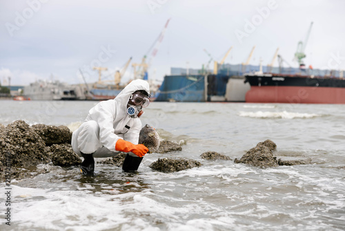 Worker in hazmat suit samples polluted seawater on a rocky shore near industrial port ships, illustrating oil spill cleanup and environmental monitoring.