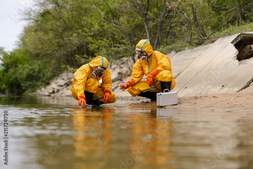 Two technicians in yellow hazmat suits sample river water near a concrete outfall, testing for pollution with protective gear and a portable case.