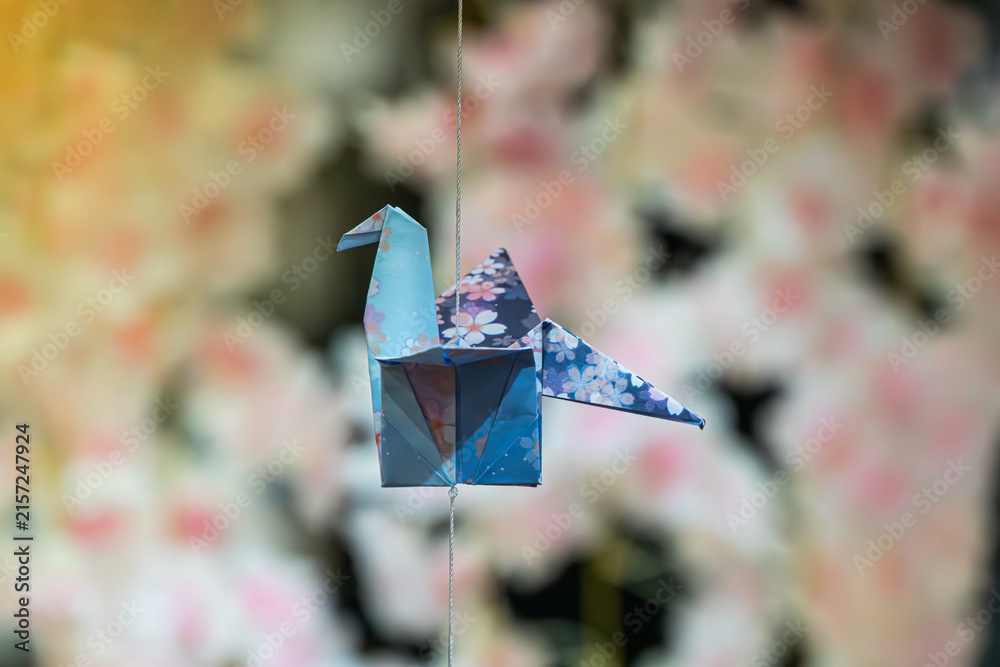 Naklejka premium Origami crane suspended by wire against floral background, intricate folded paper bird, light blue and dark blue with subtle floral pattern, soft pastel colors, gentle diffused lighting, elegant