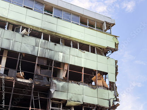 Commercial building after fire damage, partial demolition visible, light green metal panels damaged, rusted framework exposed, clear blue sky background, muted earth tones, realistic documentary