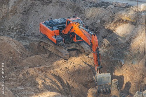 Excavator engaged in earth-moving operations within a large excavation pit, featuring loose soil and debris. Bright orange excavator with extended arm and bucket covered in dirt, highlighting dynamic