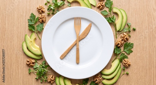 Intermittent fasting and healthy nutrition concept showing an empty white plate with wooden clock hand cutlery surrounded by keto ingredients