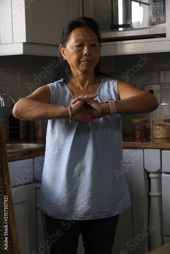 Fotografía Senior Asian woman standing at kitchen counter, clasping hands, showing smartwat