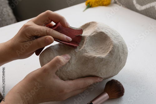 Close-up of hands shaping a plaster skull on a studio craft table, sanding with a red block and brush under soft natural light, highlighting handmade Day of the Dead sculpture prep.