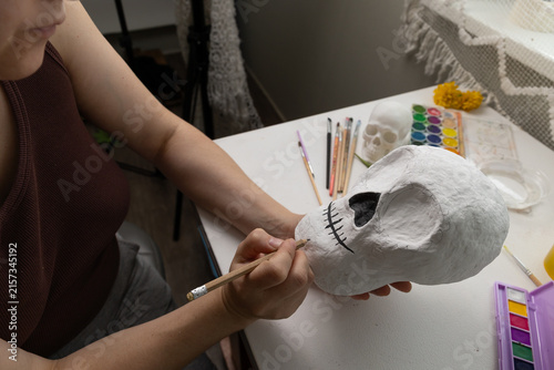 Photography Close-up of a handmade Day of the Dead skull in progress on a white craft table, with pencil sketch lines, left-hand support, and a small palette, brushes, a yellow flower, and crochet blanket
