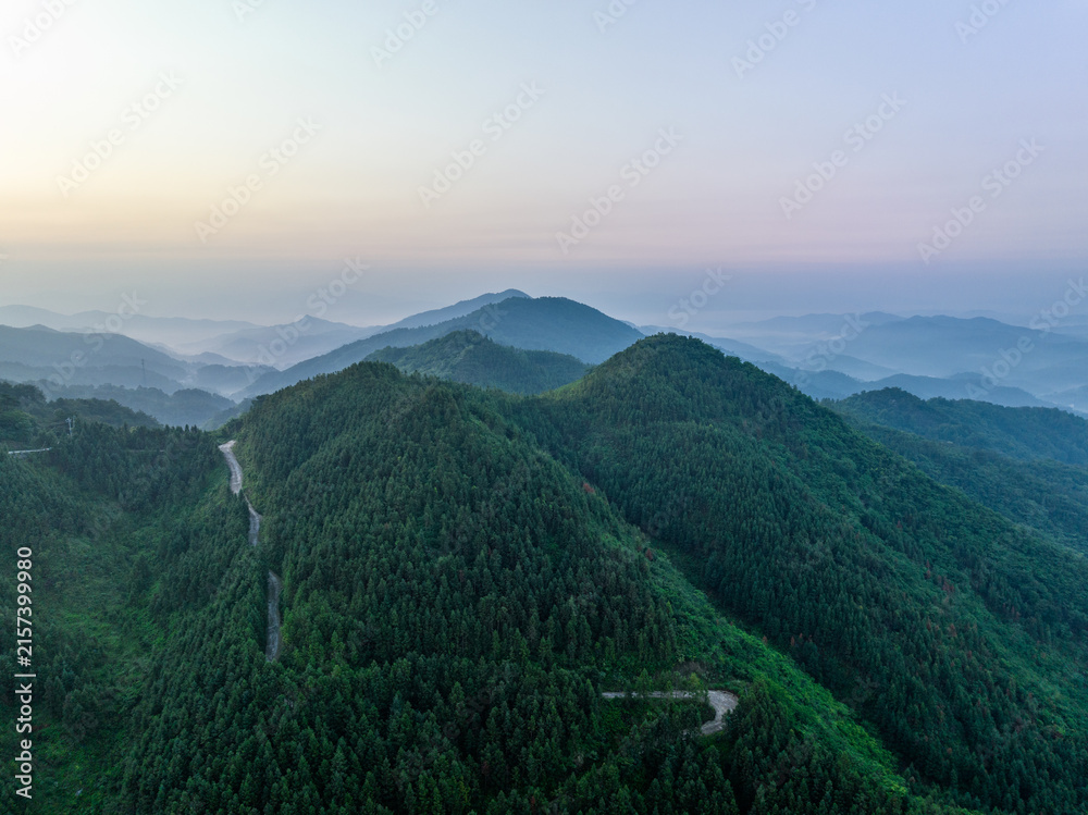 Fototapeta premium Layered forested mountain ranges with winding roads, shrouded in soft morning mist under a pale dawn sky.