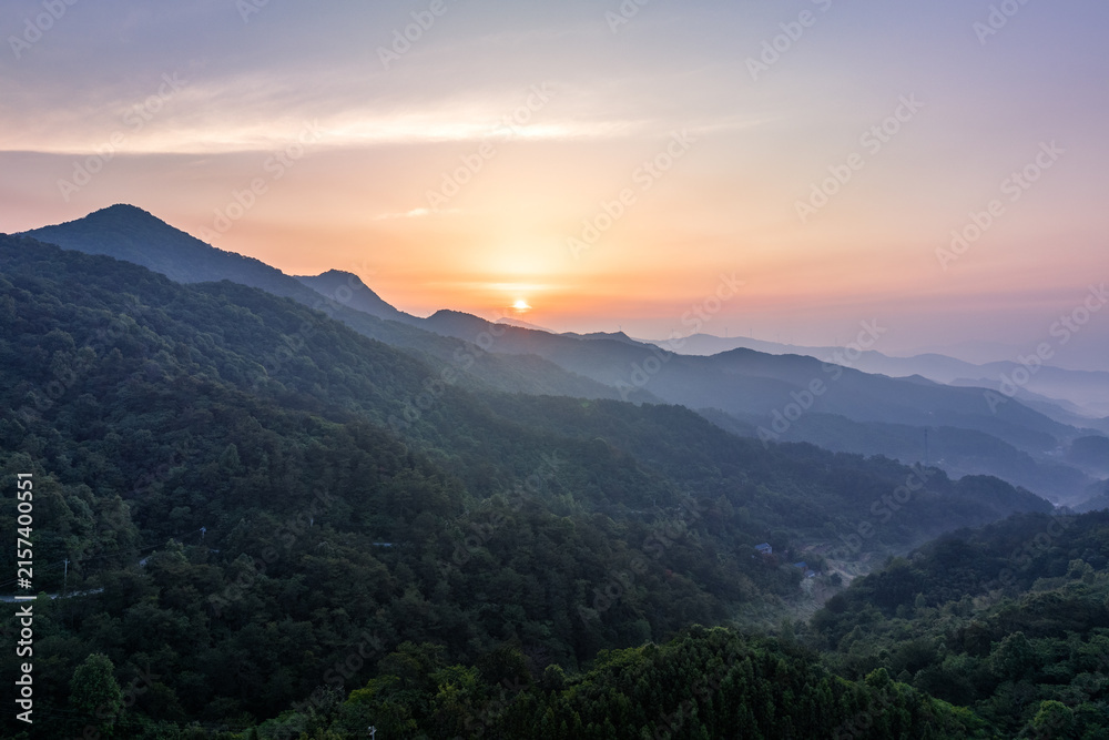 Fototapeta premium A serene sunrise illuminates layered forested mountain ranges with soft mist in the valleys and warm sky hues.