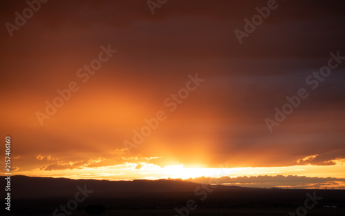 Canvas Print Dramatic fiery orange sunset with sunbeams and crepuscular rays bursting upward behind dark stormy clouds over a silhouetted mountain horizon