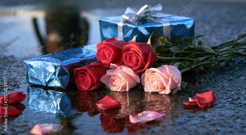 Two blue gifts with ribbon next to red and pink roses on wet ground