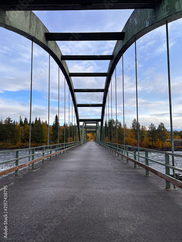 Symmetrical view along a steel arch bridge crossing a river, surrounded by colorful autumn forest under a cloudy blue sky in northern Sweden.
