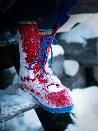 Close-up of a traditional Sámi winter boot covered with fresh snow, featuring bright red fabric, blue laces and decorative woven trim.
