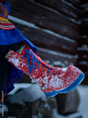 Close-up of a traditional Sámi winter boot covered with fresh snow, featuring bright red fabric, blue laces and decorative woven trim.