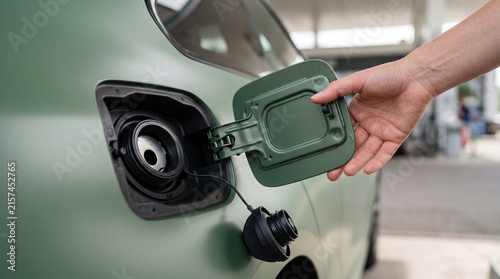 A hand is shown opening the fuel tank door of a matte green vehicle, revealing the open gas cap ready for refueling at a petrol station.