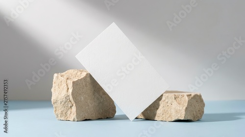 Minimalist scene with two rough stones and a white card against a light blue background with diagonal light and shadow.