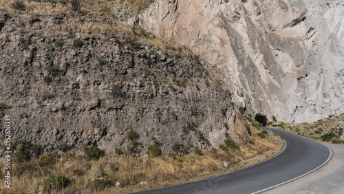 A high-altitude highway bends around a cliff and goes into a dark tunnel. Yellow grass, bushes on the side of the road. A warning road sign.  Rocky slopes of the cliffs. Peru