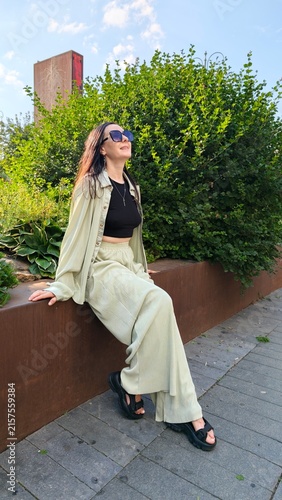 A young woman sits on a bench in a park wearing a matching outfit. She has sunglasses on and smiles as she enjoys the sunny day surrounded by greenery