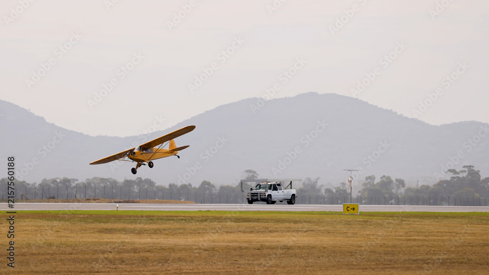 Fototapeta premium Yellow Vintage Piper Cub Aircraft Taking Off Beside a White Truck