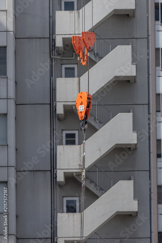 Construction crane pulley system with orange hook block in front of a grey urban building's zigzagging concrete emergency staircase in Japan.