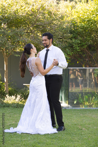 Couple dancing on lawn, bride in white lace gown train, groom black tie, glass railing reflecting