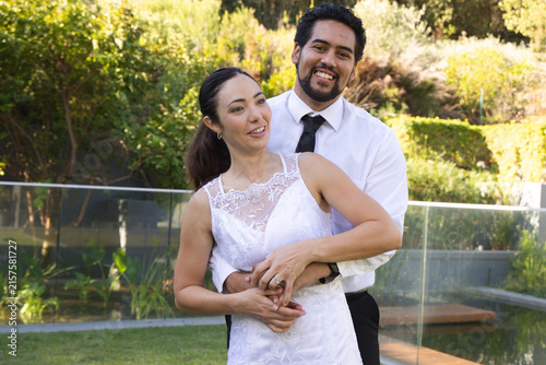 Diverse couple embracing in yard by glass railing wearing white-lace dress and black tie with rings