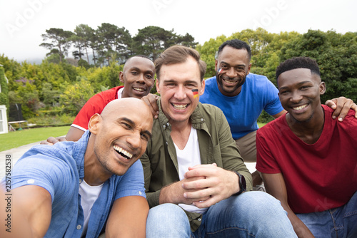 Group of five men leaning in smiling while holding smartphone on wooden deck, showing face paint