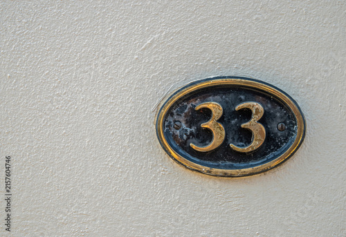 Weathered oval metal house number 33 plaque mounted on a textured white exterior wall.