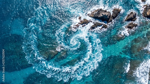 Aerial top down view of ocean waves swirling around jagged coastal rocks