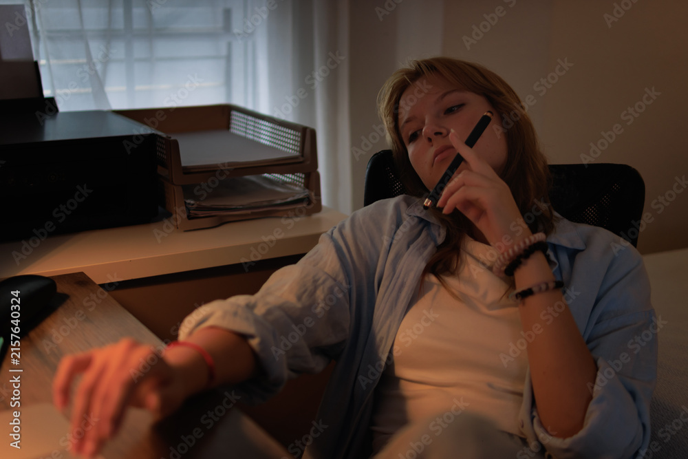 Fototapeta premium Young female student feeling bored and tired during late night study, procrastinating on homework or school project in a dimly lit home office